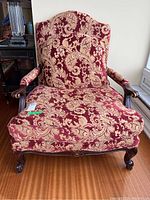 Front view of antique style upholstered armchair with red and cream floral damask fabric and wooden carved frame and armrests.
