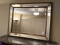 Front view of rectangular beveled mirror with gold beaded trim frame, positioned on dark marble surface, reflecting ceiling light fixture and door.