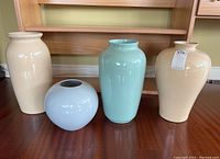 Four tall pastel ceramic vases on wooden floor near a wall, showing their different shapes and soft colors.