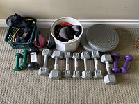Top down view showing the variety of dumbbells (hex and coated), kettlebells, resistance bands in basket, ankle/wrist weights in bucket, and a gray balance disc.