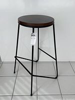 Black wrought iron bar stool front view showing round wooden seat and angled legs on tiled floor.