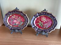 Front view of both ceramic floral plates displayed on metal stands, showing detailed paintwork and edge shape.