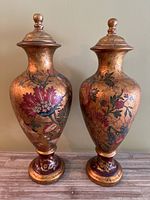 Front view of two tall ceramic urns with bronze finish and red floral patterns on wooden surface against beige wall.