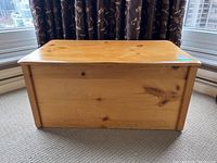 Front view of pine blanket box showing natural wood grain and knots, on carpeted floor with curtains in background.