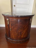 Front view of round end table showing rich mahogany veneer with flamed grain and decorative brass/bronze hardware on door and gilded leaf carvings.