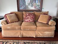 Front view of tan velvet three-seat sofa with five decorative pillows in coordinating colors placed along backrest. Sofa has a pleated skirt, plush cushions, and rolled armrests.