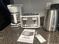 White Toastmaster drip coffee maker, stainless steel Toastmaster toaster, and Oster electric kettle placed on countertop. Instruction manual for toaster visible.