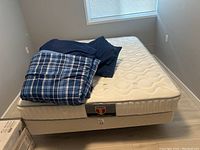Metal queen size bed frame mattress covered with blue checkered comforter and navy blue bedding, placed against corner of room with wooden floor and window.