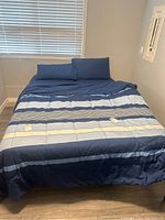 Bed made with navy blue striped comforter and two navy blue pillows in front of a window with blinds.