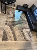View of textured shag carpet with curved striped pattern in beige, gray and brown tones on wood floor with glass coffee table and black furniture.