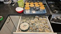Drawer open showing a set of plates, mugs, shakers, glassware and ten glass jars with cork lids arranged in a tray.