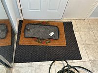 View showing three floor mats on the floor near a door: a large black rubber mat, a brown coir mat with floral wreath, and a smaller black rubber tray mat on top.