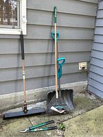 Photo of set of gardening tools including lawn rake, snow shovel, broom and hedge shears leaning against gray wall outdoors