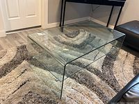 Glass coffee table placed on a multi-toned rug, showing its bent glass construction and smooth edges from a top corner angle.