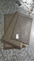 Three mats stacked on a tiled floor, including two foam mats and one rubber-backed 'wipe your paws' mat.