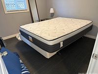 Photo showing side angle of queen mattress and metal bed frame with wheels under a window.