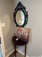 Photo showing black ornate oval mirror on wall above dark semi-circle wooden table with picture leaning on table.