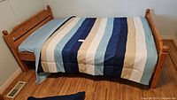Full view of pine bed frame with foam mattress covered by blue, beige, and white striped bedspread.