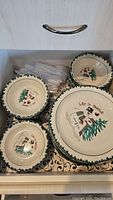 Photo showing various sized winter themed bowls with a snowman, pine tree, and 'Let it Snow' artwork in green rimmed ceramic design stored in a drawer with disposable forks.