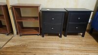 Two matching black wooden side tables with two drawers each and one brown rolling cart with two shelves on casters, all placed on wooden floor showing scratches and wear.