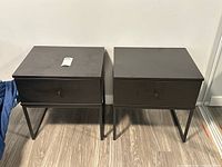 Front view of two matching black wood side tables with single drawer and metal legs side by side on wood floor.