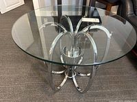 Side-angle view showing the round clear glass top and the sculptural curved metal base of the table on a carpeted floor.