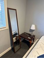 Full-length mirror leaning against wall beside wooden side table with a table lamp on top, next to a bed.