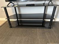 Full frontal view of the black TV stand showing top glass shelf, middle glass storage shelf, and bottom solid shelf, standing on carpeted floor.