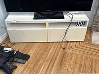 Front view of white wooden TV stand with TV on top and three white cabinet doors, two open shelves, and several cables and remote controls on the floor in front.
