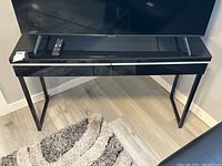 Full view of the black TV stand console table with glass top, two drawers, and metal legs, shown with a remote on top and part of a TV screen visible above.