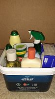Top view of assorted personal care and cleaning items in a white JetBlue labeled plastic container, featuring spray bottles, baby powder, lotion, and wipes.