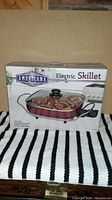 Box view of red Americana Classics electric skillet with glass lid, showing cooking surface with food and power cord.