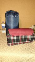 Blue sleeping bag compressed in nylon storage sack next to a water bottle for size comparison, folded plaid blanket in brown and red carry case on a beige textured surface.