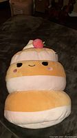 Front view of Squishmallow plush toy resembling a layered cake with a strawberry on top, showing face and colors.