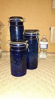 Three cobalt blue glass storage jars with metal clamp lids, arranged from smallest to largest beside a water bottle for size comparison, showing used condition and some rust on clamps.