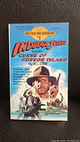 Front cover of 'Indiana Jones and the Curse of Horror Island' paperback book showing illustrated cover artwork with Indiana Jones and title details