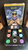 Photo showing the Marvel Splendor board game box with Thanos holding the Infinity Gauntlet and Marvel heroes in the background, open display of tokens and cards inside the plastic tray