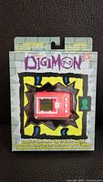 Front view of sealed Digimon 20th Anniversary virtual pet game in packaging showing the red virtual pet device inside.
