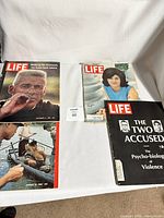 Photo shows four LIFE magazines arranged flat on a white cloth. Covers include a close-up of a man smoking, a woman in blue, a social issue black and white cover, and a Vietnamese war scene with soldiers in water rescue
