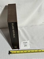 Side view of the Folio Society Plato Republic book showing black hardcover spine with gold text and Folio Society logo; brown slipcover beside it, with measuring tape showing approx. 12 inches tall