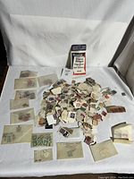 Photo showing multiple small envelopes and loose piles containing used postage stamps of various kinds and colors, along with a pack of 1000 Stamp Hinges.