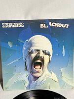 Front cover of Scorpions Blackout album showcasing screaming man with glasses and headband in front of a shattered glass design.