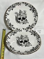 Set of four white ceramic dinner plates with black skull and floral design and butterfly detail in the center and floral border around rim. Photograph taken with measuring tape showing plate diameter about 27 cm.