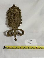 Front view of the vintage brass wall mounted coat hook showing detailed Art Nouveau floral decoration and double curved hook with scrolls. A tape measure for scale shows approximately 7 inches in height and 4 inches wide at the widest point.
