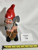 Frontansicht des Vintage Gartenzwergs mit roter Mütze und Hacke, auf weißem Hintergrund mit Maßband als Größenreferenz von ca. 20 cm Höhe.