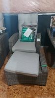 Front view of wicker-look chair and ottoman with gray cushions and included weather cover in packaging