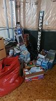 Photo of assorted Christmas decorations including snowman figurines, mugs, gift boxes stacked near a wall, and a vertical 'Happy Holidays' sign.