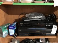Shelf showing Panasonic DVD/CD player DVD-RV32 on top, JVC VHS Hi-Fi stereo VCR HR-JJ698U below, with boxes of blank CDs to the side and remote control placed on top of DVD player.