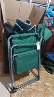 Folded green camp chair with metal frame and cooler table stored against wall with cardboard box behind