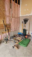 Various garden tools and turf remnants placed on the garage floor, showing a grouping of shovels, rakes, and wire tomato cages beside a green turf remnant roll.